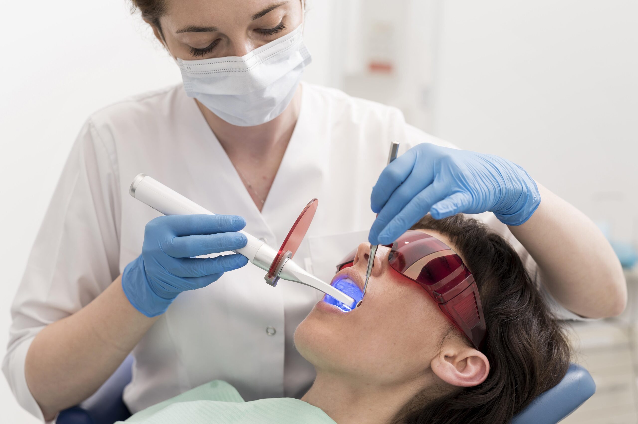 female-patient-having-procedure-done-dentist A dentist using a dental laser to whiten teeth, with the patient wearing protective orange glasses.