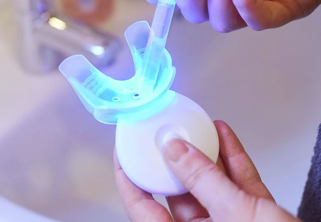 A hand holding a small device with a glowing blue light, likely used for at-home teeth whitening.