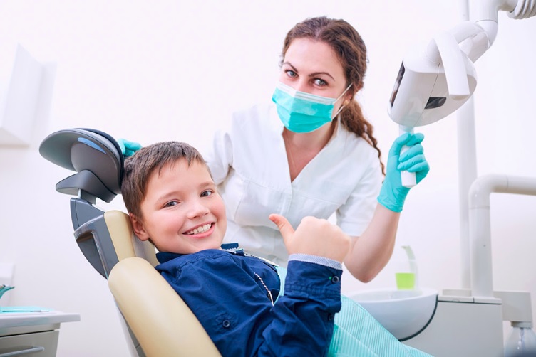 A young boy sitting in a dental chair with a dentist, who is holding a dental tool and smiling.