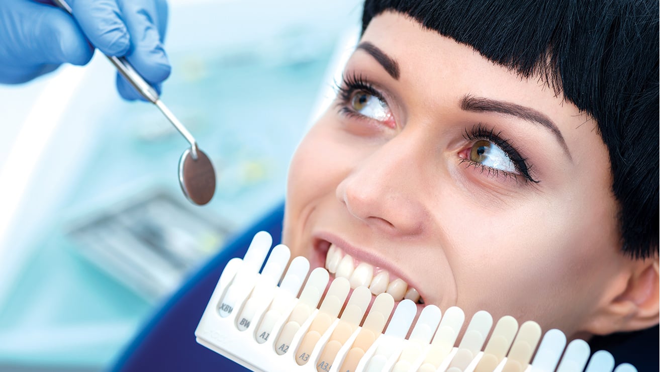 professional_dentist_whitening طبيبة أسنان تستخدم مرآة صغيرة لفحص الأسنان، بينما تبتسم المريضة وتحمل لوحة ألوان الأسنان