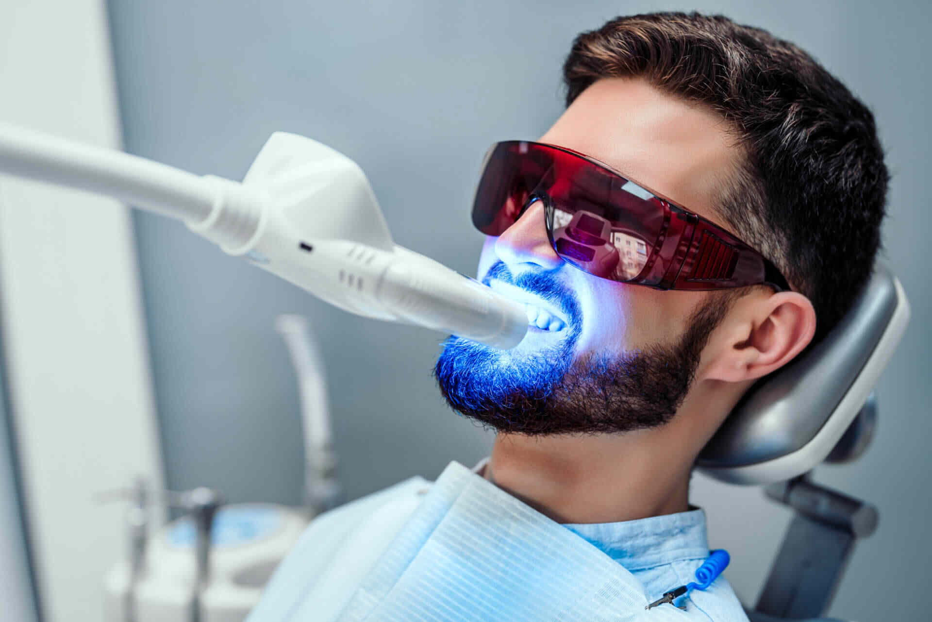 Nano Teeth Whitening Man undergoing professional laser teeth whitening treatment at a dental clinic, wearing protective red glasses and sitting in a dental chair.