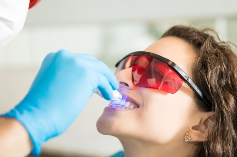 Dentist performing nano teeth whitening using a handheld LED light while the patient wears protective red glasses.
