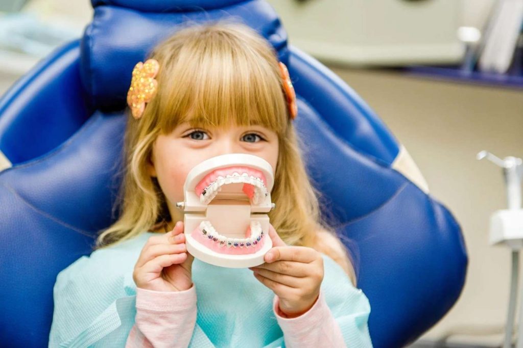 Young girl sitting in a dental chair holding a model of teeth with colorful braces during a pediatric dental visit.
